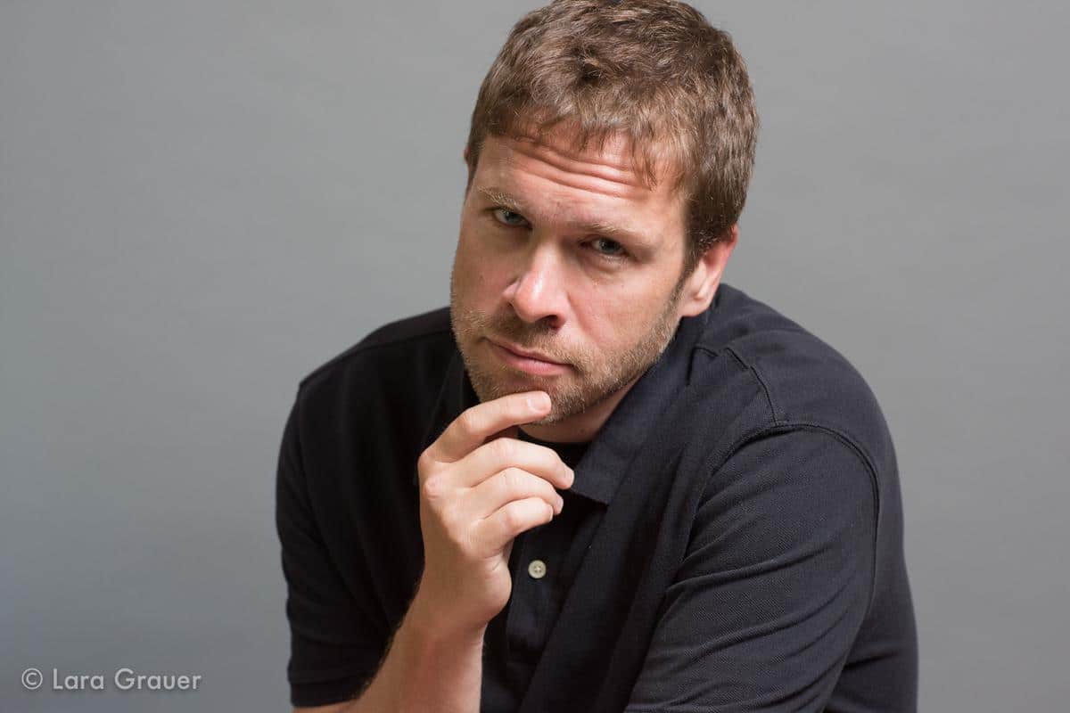 man in a black polo shirt looking curious with his hand on his chin.