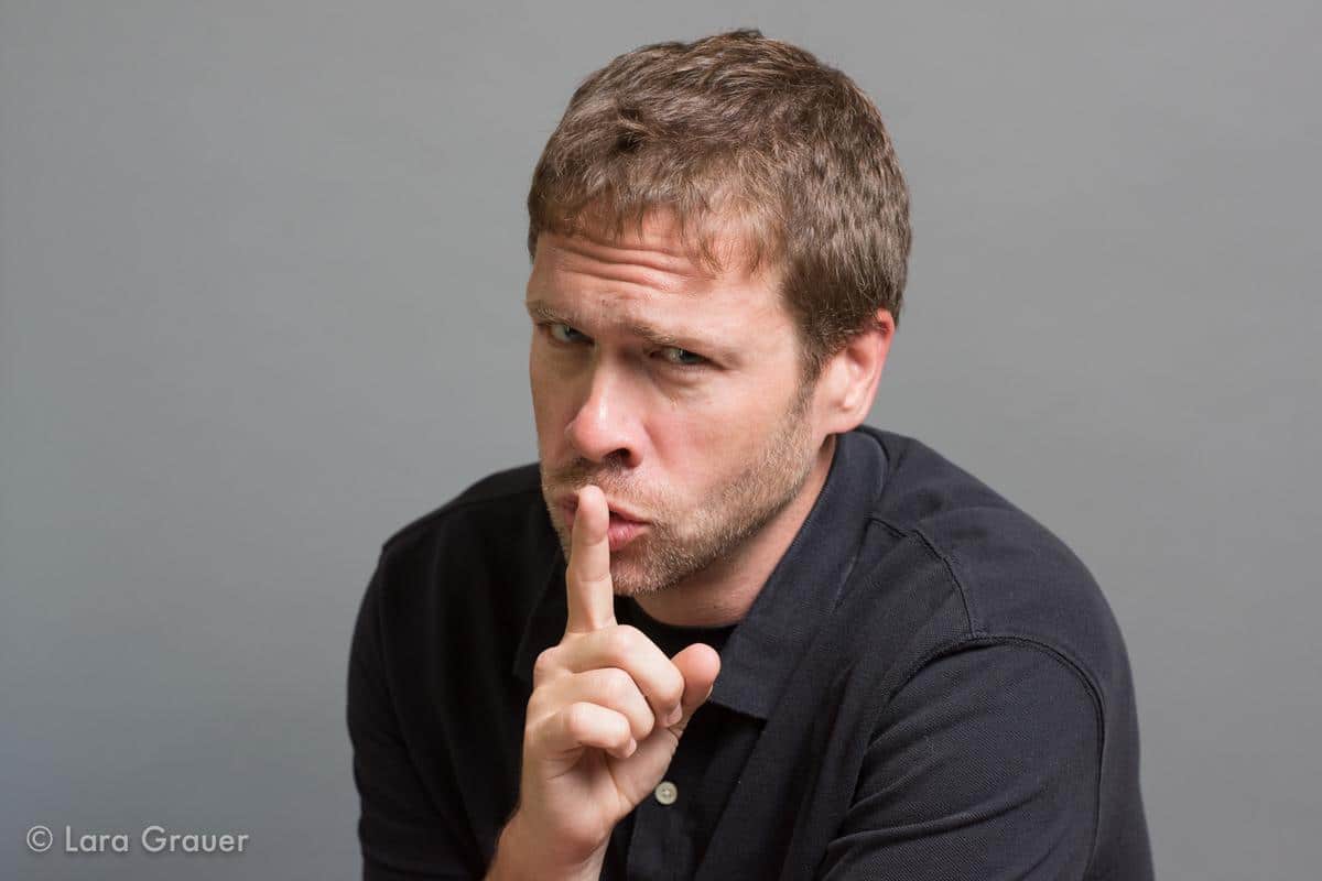 man in a black polo shirt making a shush expression with his finger on his mouth.