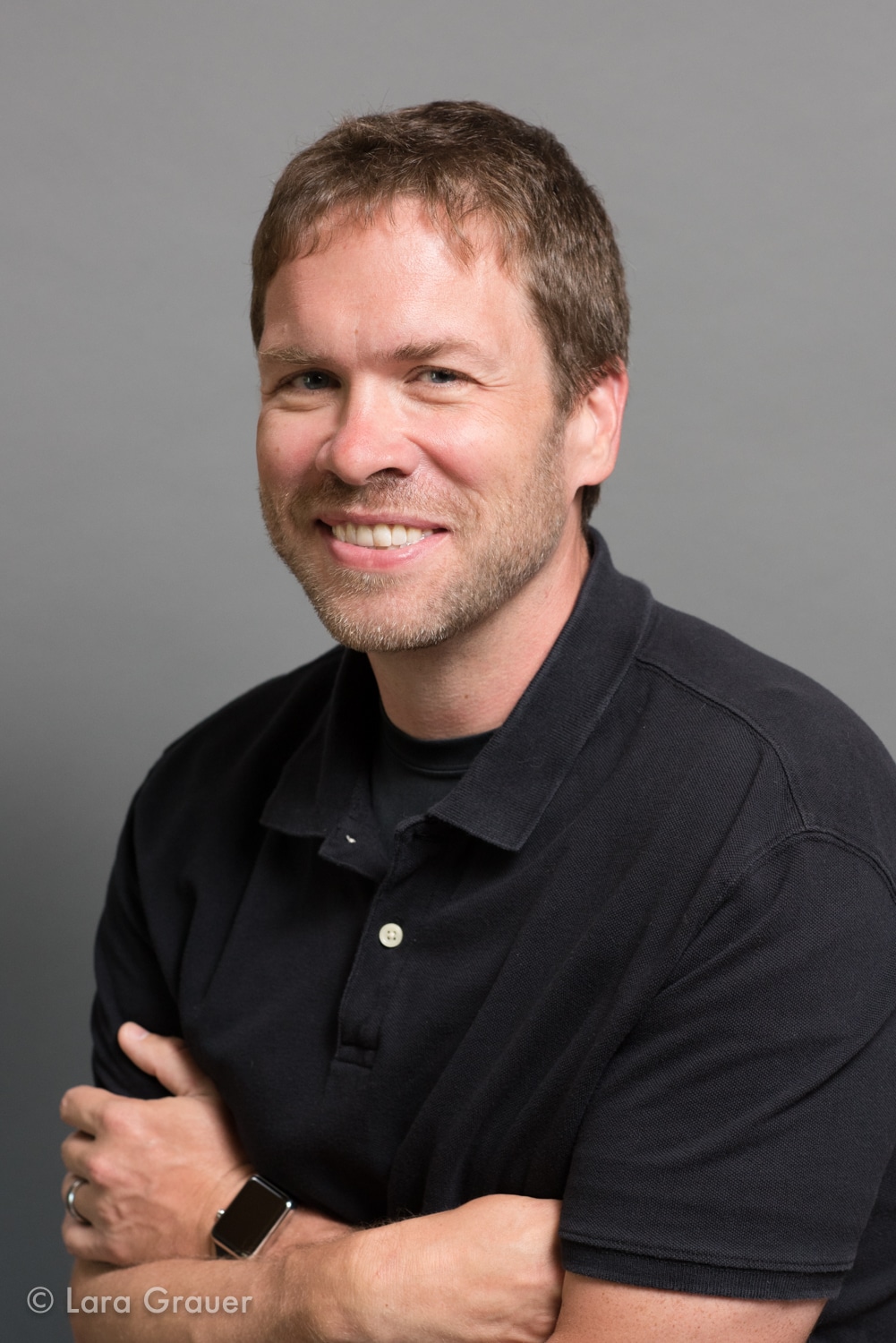 man in black polo shirt smiling and crossing his arms