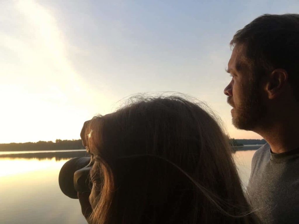 women taking a picture of a lake while a man standing next to her stares off into the distance