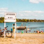 beach waterfront with people swimming and a bike leaning on a sign saying lifeguard on duty.