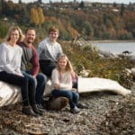 mom, dad, son, and daughter sitting on a log on a waterfront for a family photo.