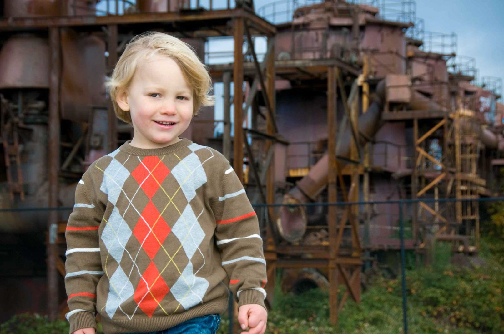 portrait of a young boy in an argyle sweater in front of abandoned buildings.