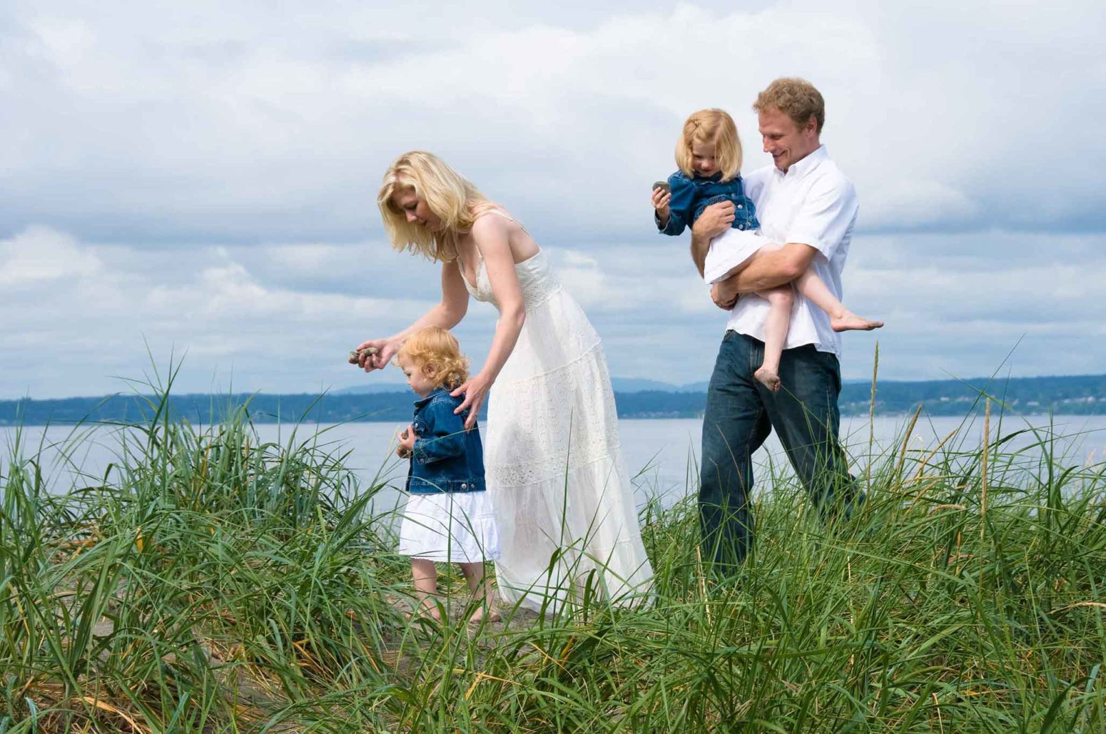 family of four walking through the grass at the beach with a waterfront in the background.