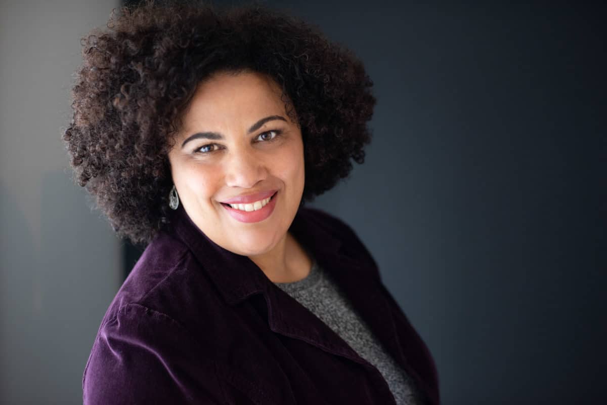 headshot of a woman with dark brown curly hair and purple jacket facing right and looking at camera