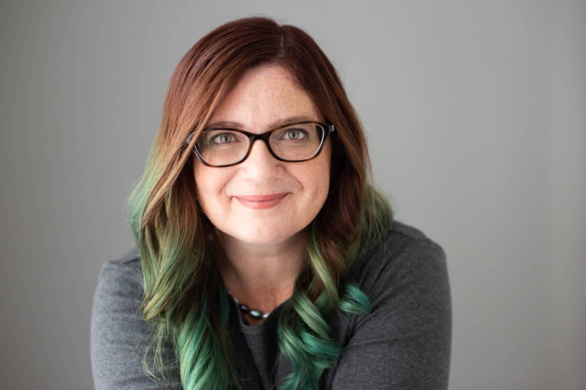 woman with glasses and brown hair with green highlights smiling towards camera