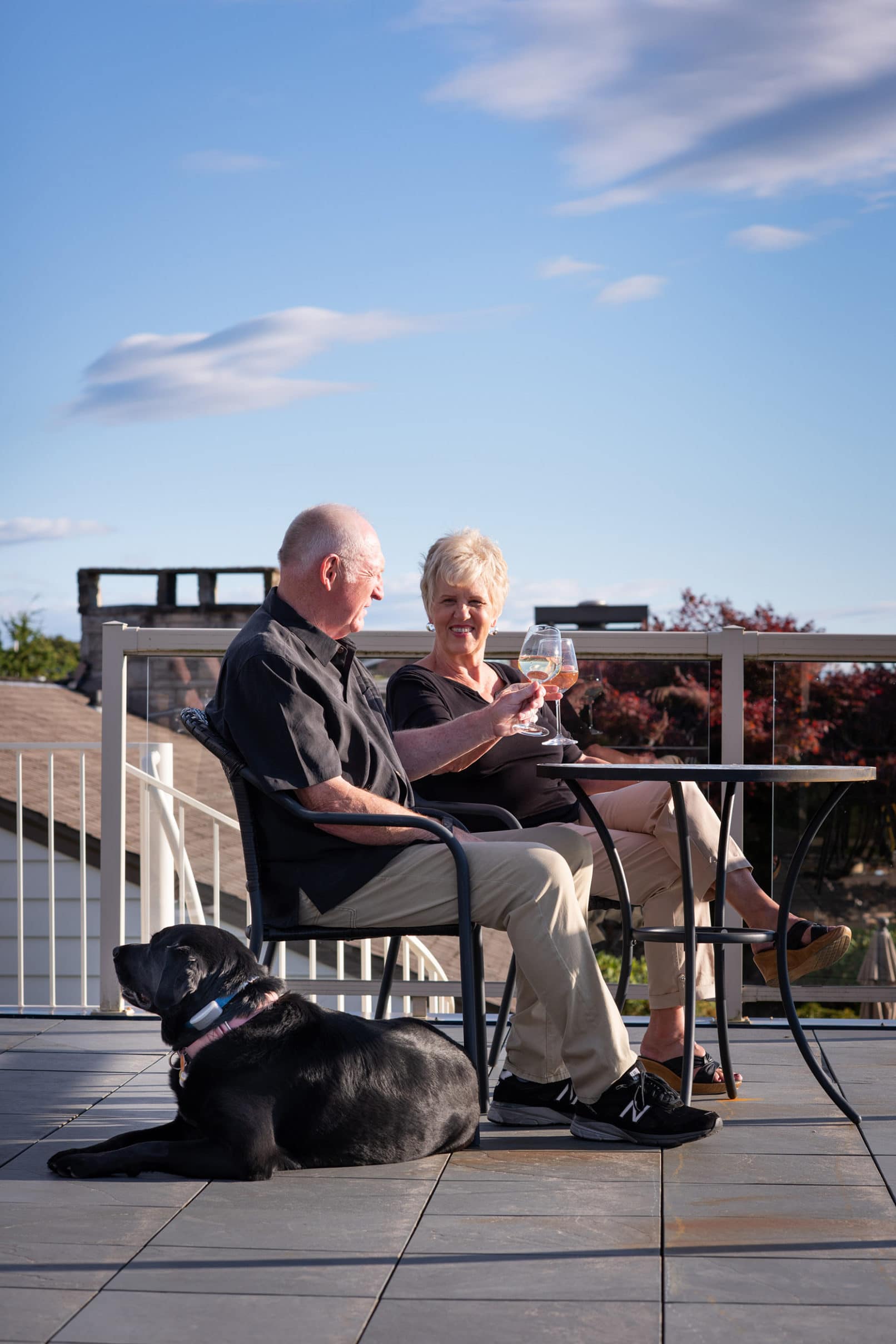 photo of an elderly couple having wine outside with their black dog nearby