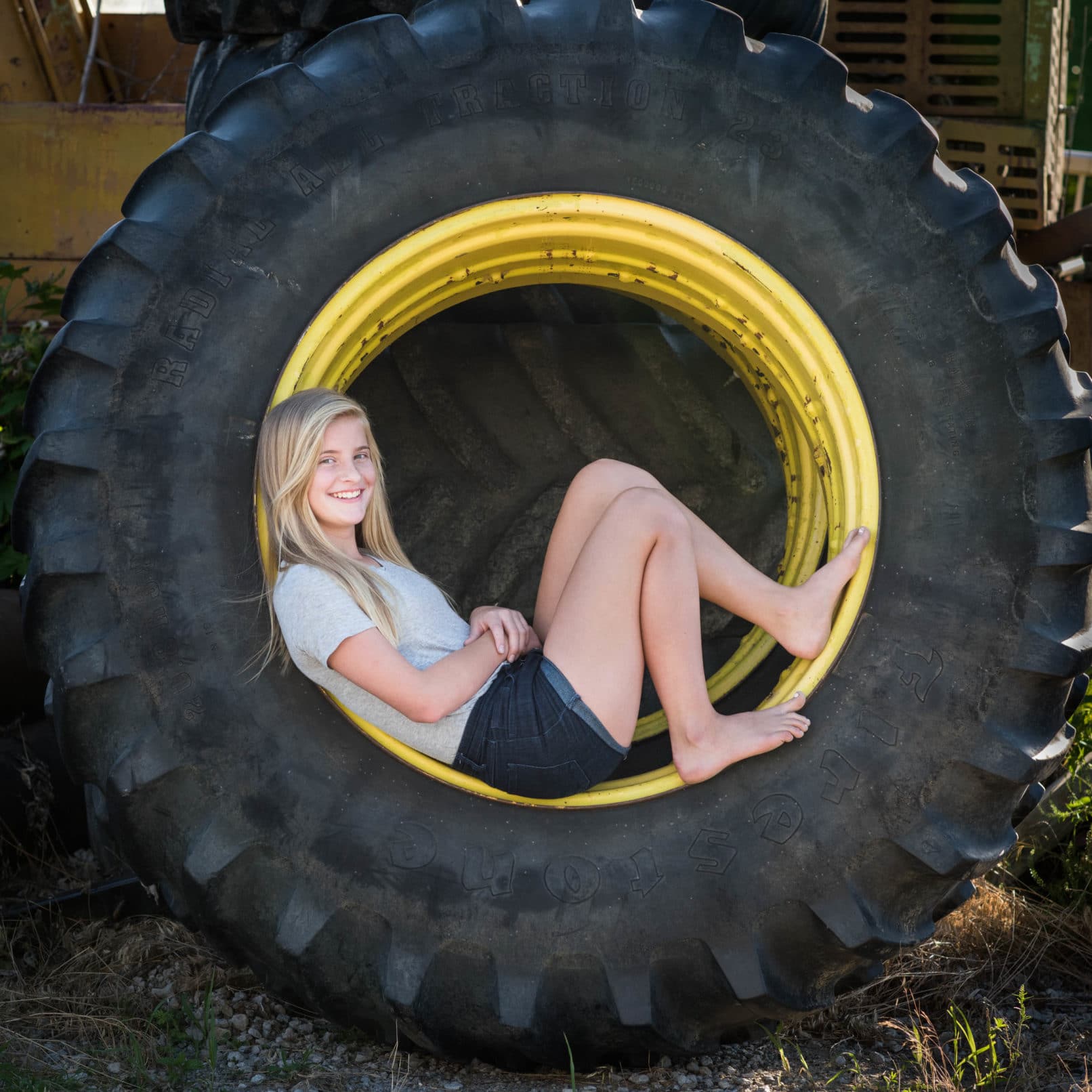 young blonde girl sitting inside of a large tire with barn as background.