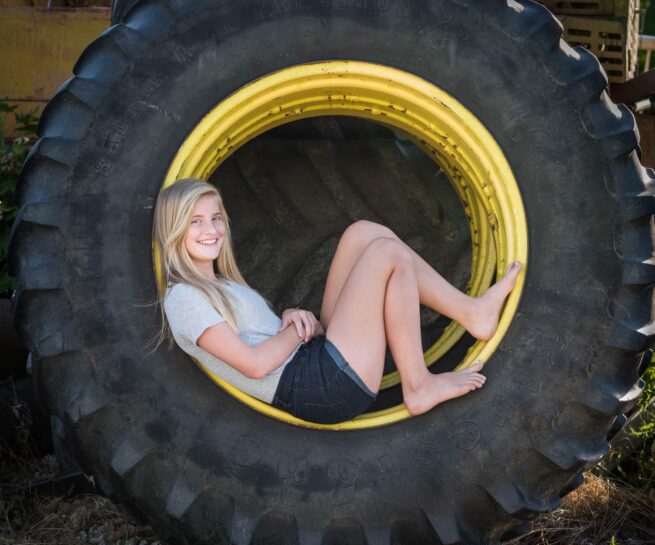 young blonde girl sitting inside of a large tire with barn as background.