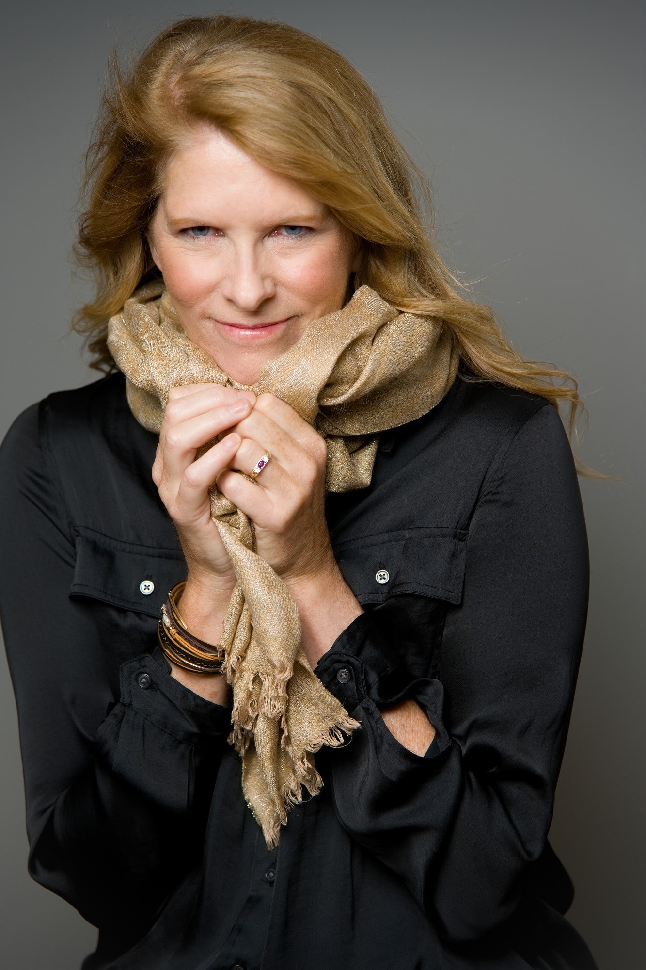 middle-aged blonde woman in black wearing a tan scarf with her hands grabbing it