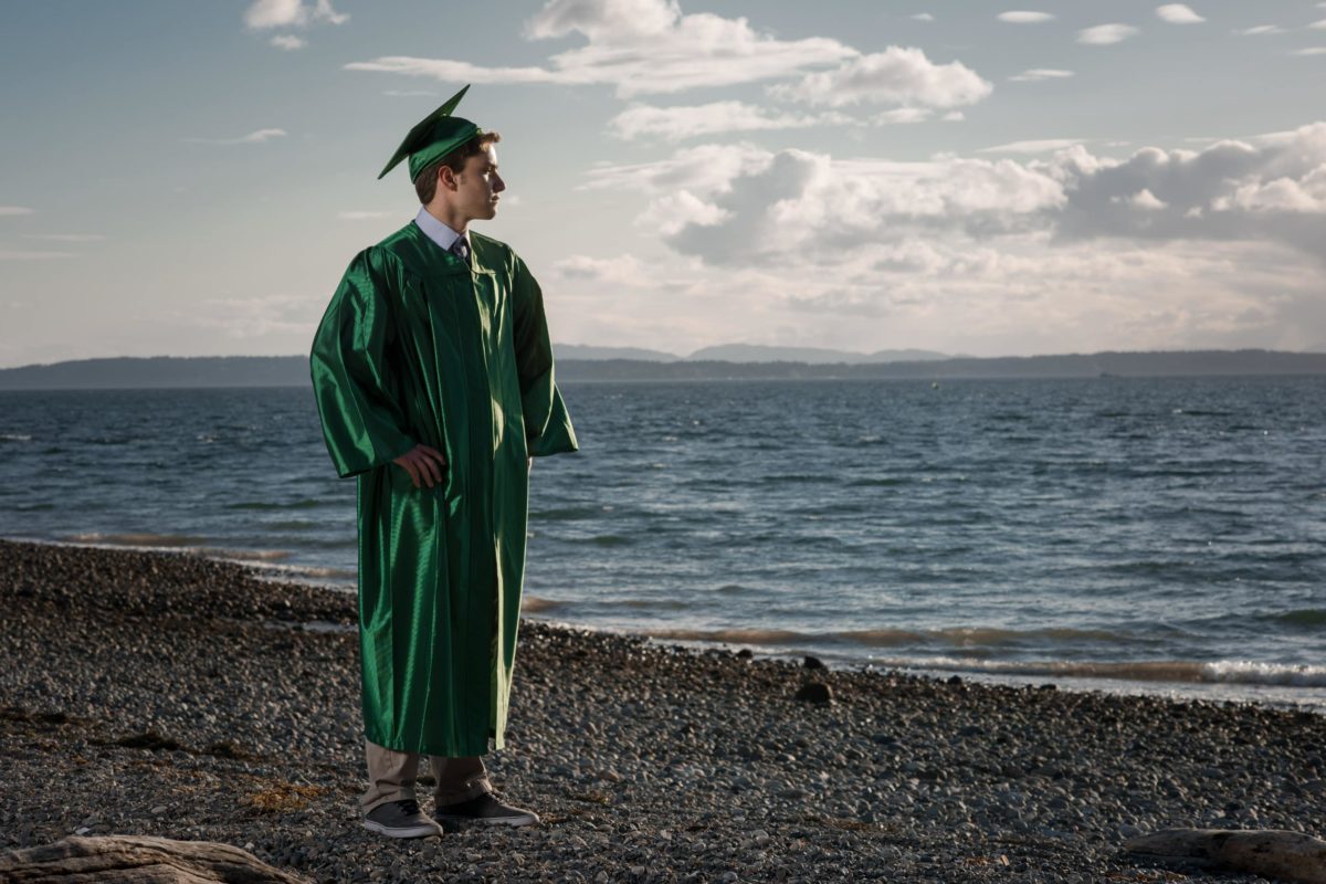 boy in a green high-school graduation gown staring off into the ocean