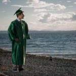 boy in a green high-school graduation gown staring off into the ocean