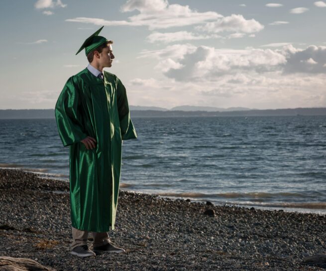 boy in a green high-school graduation gown staring off into the ocean