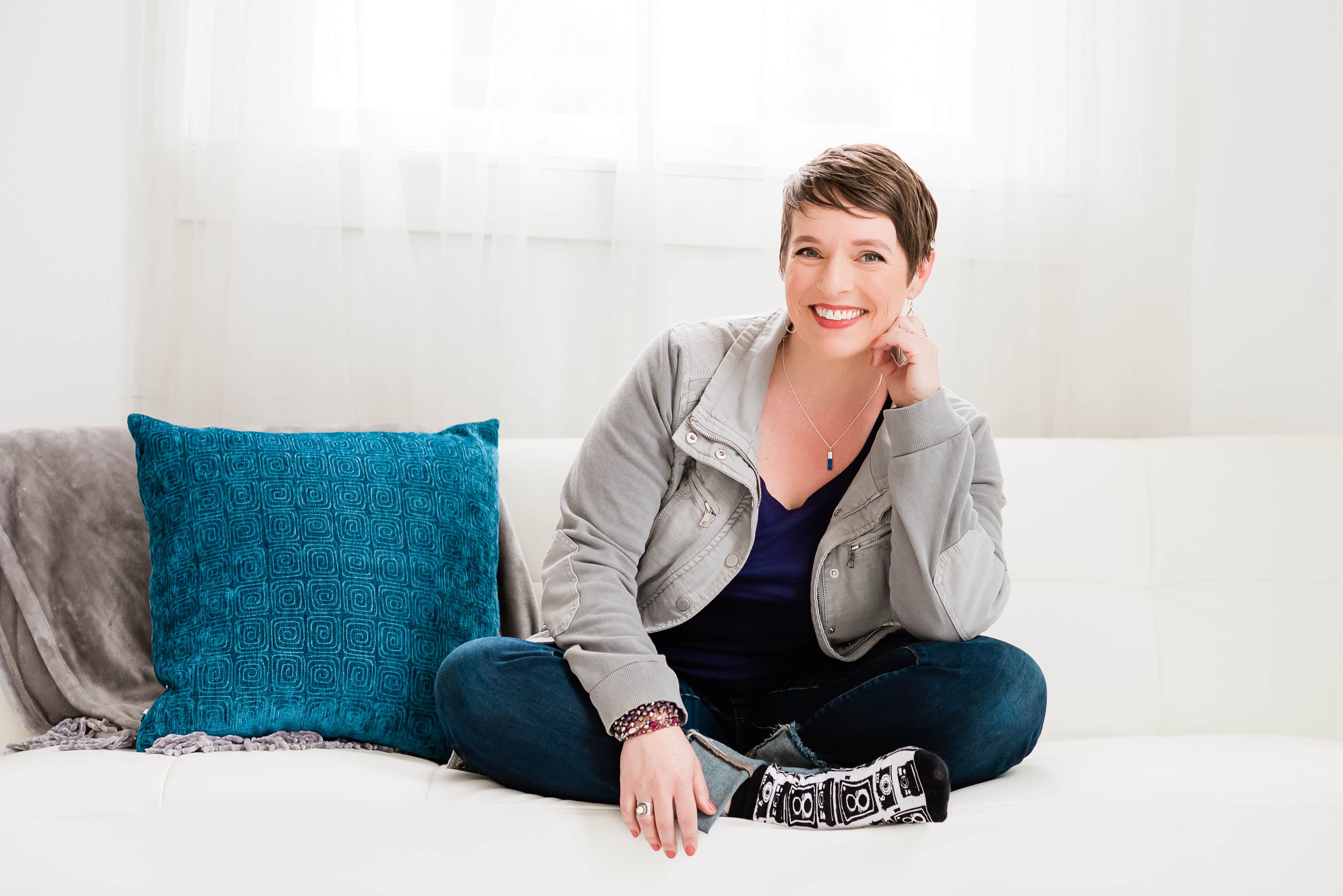 smiling woman sitting crossed leg on a white couch with a throw and pillow to her left side.