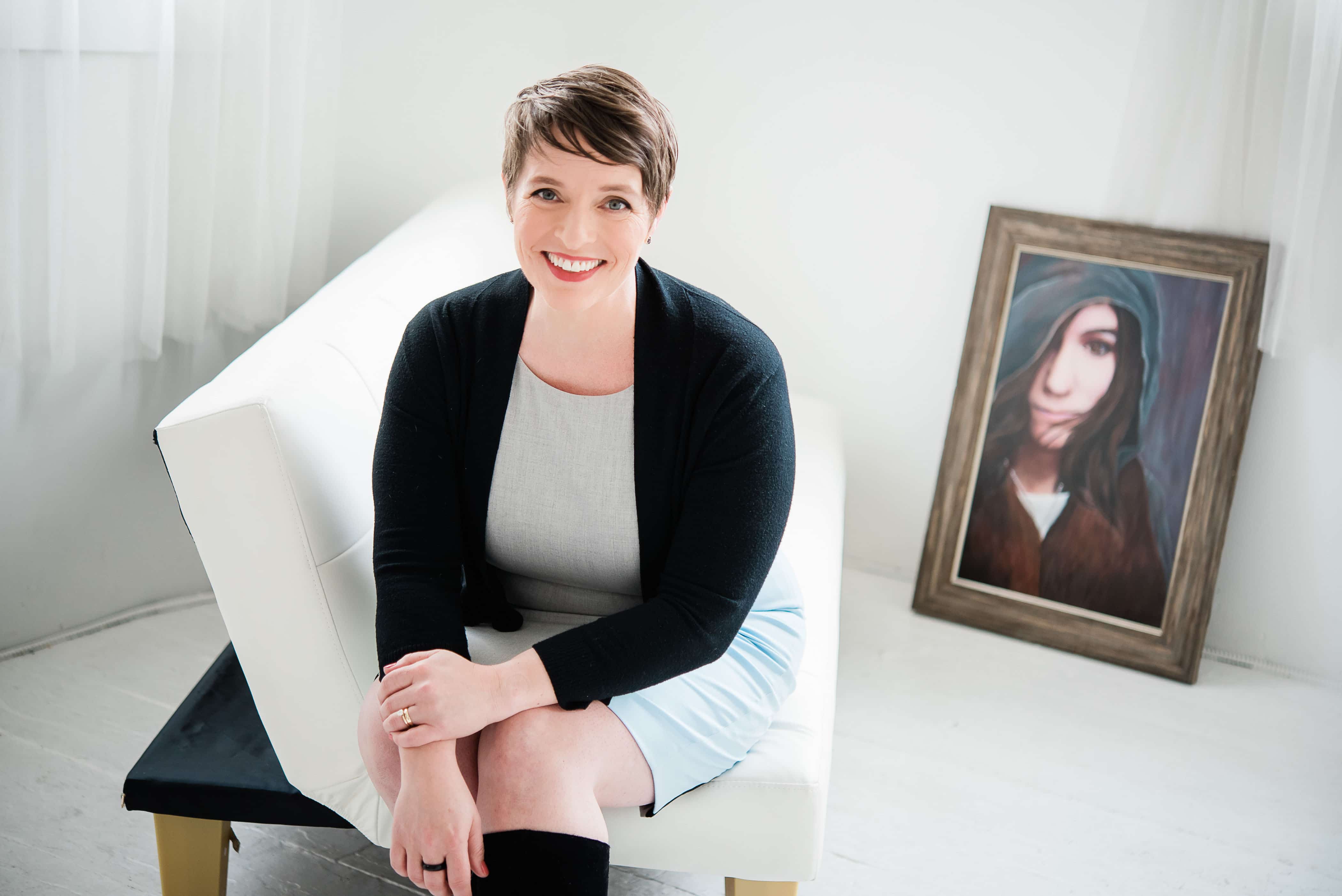 woman sitting on a white love seat with white backdrop and floor and a wall portrait leaning against wall.