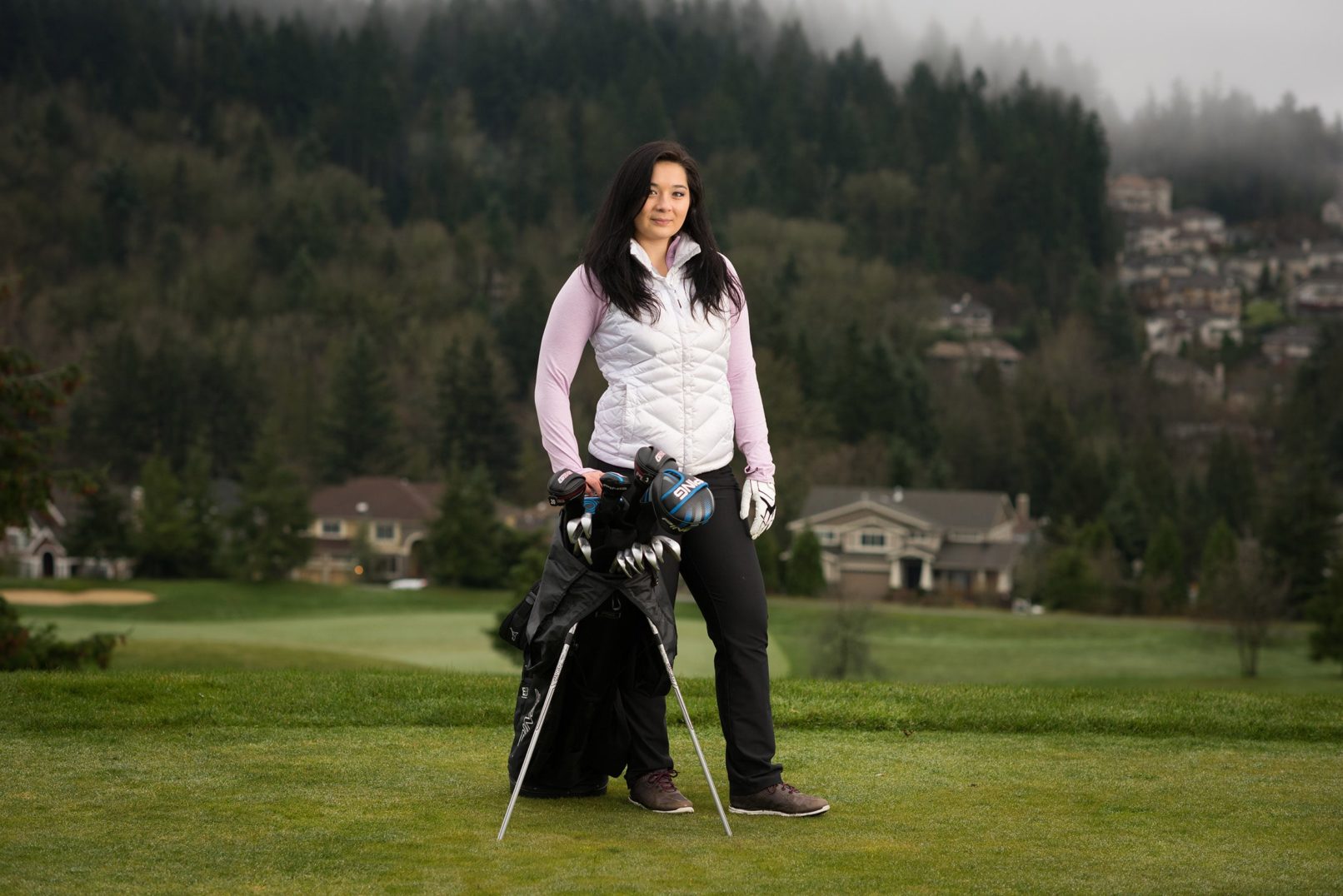 women outfitted for golf with golf clubs held up by tripod attached to golf bag and houses and hills beyond the course.