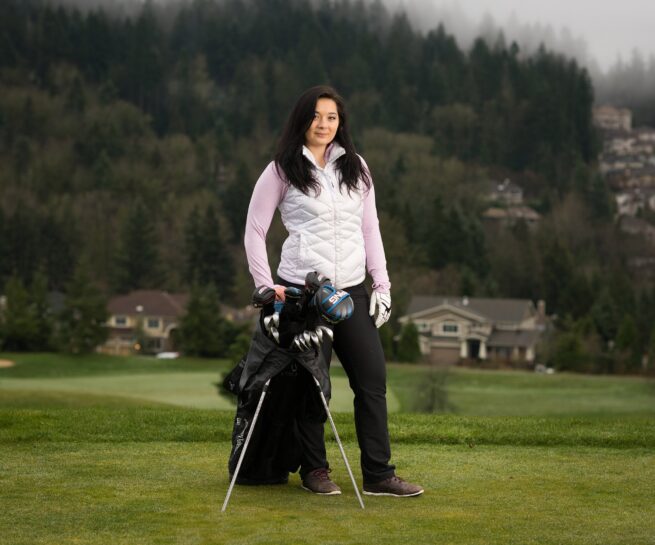 women outfitted for golf with golf clubs held up by tripod attached to golf bag and houses and hills beyond the course.