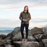 girl wearing a jacket and jeans stands on rocks with Puget Sound in the background.