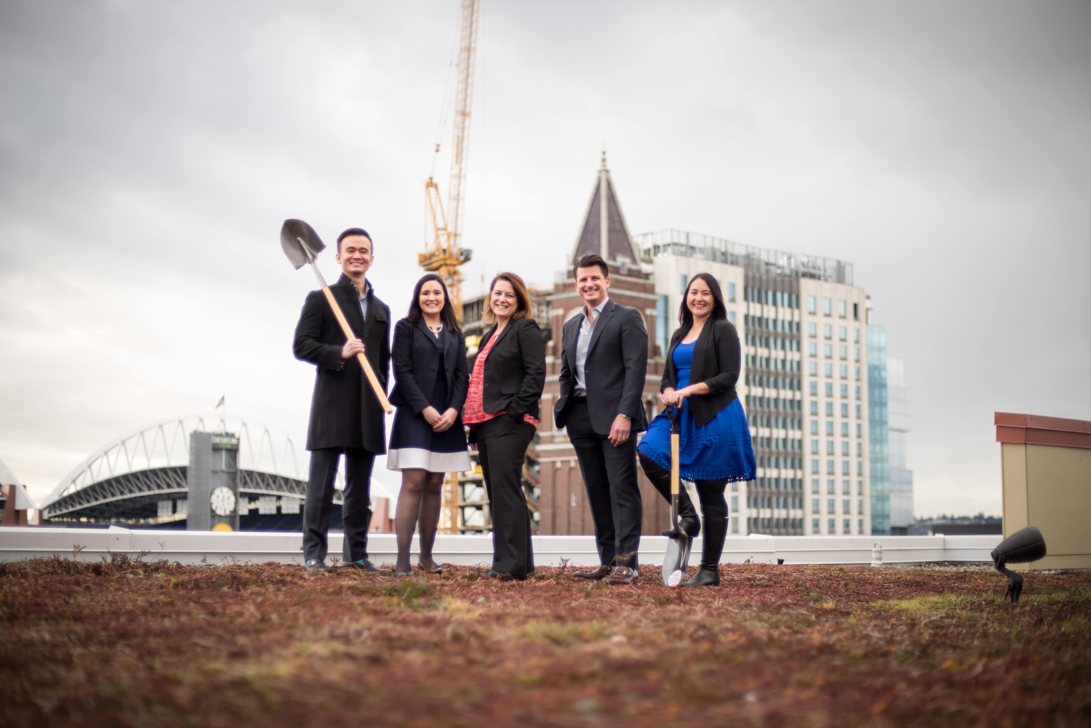 two men, one holding a shovel and three women, posing on the roof top with a crane and buildings in the background.