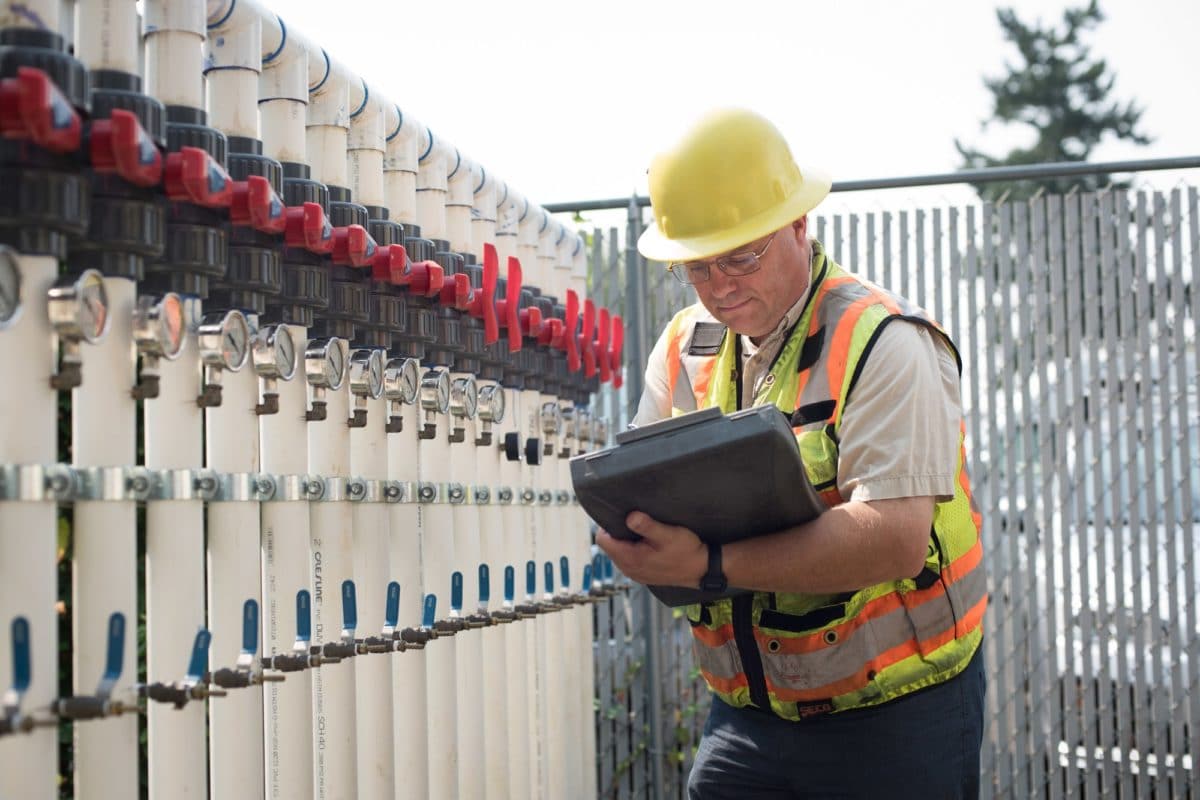 man in construction hard hat and vest inspecting clipboard while standing next to many valves