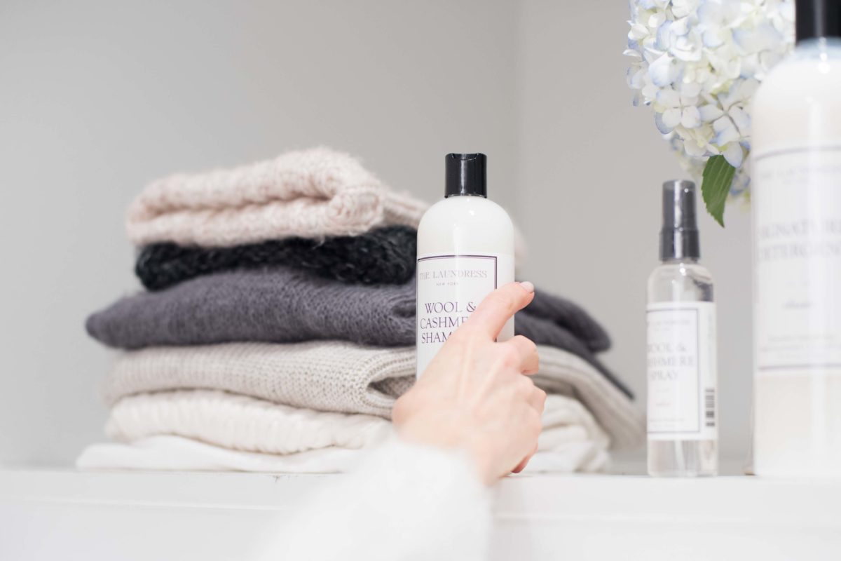 women's hand putting a cleaning product for wool and cashmere on a shelf with sweaters, flowers and two other bottled products.