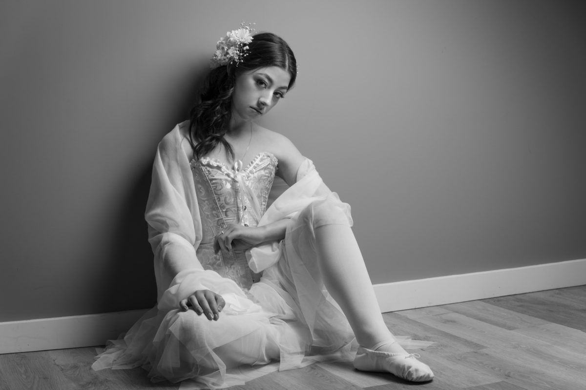 Young woman ballerina sitting with one leg crossed against a wall with wood flooring.