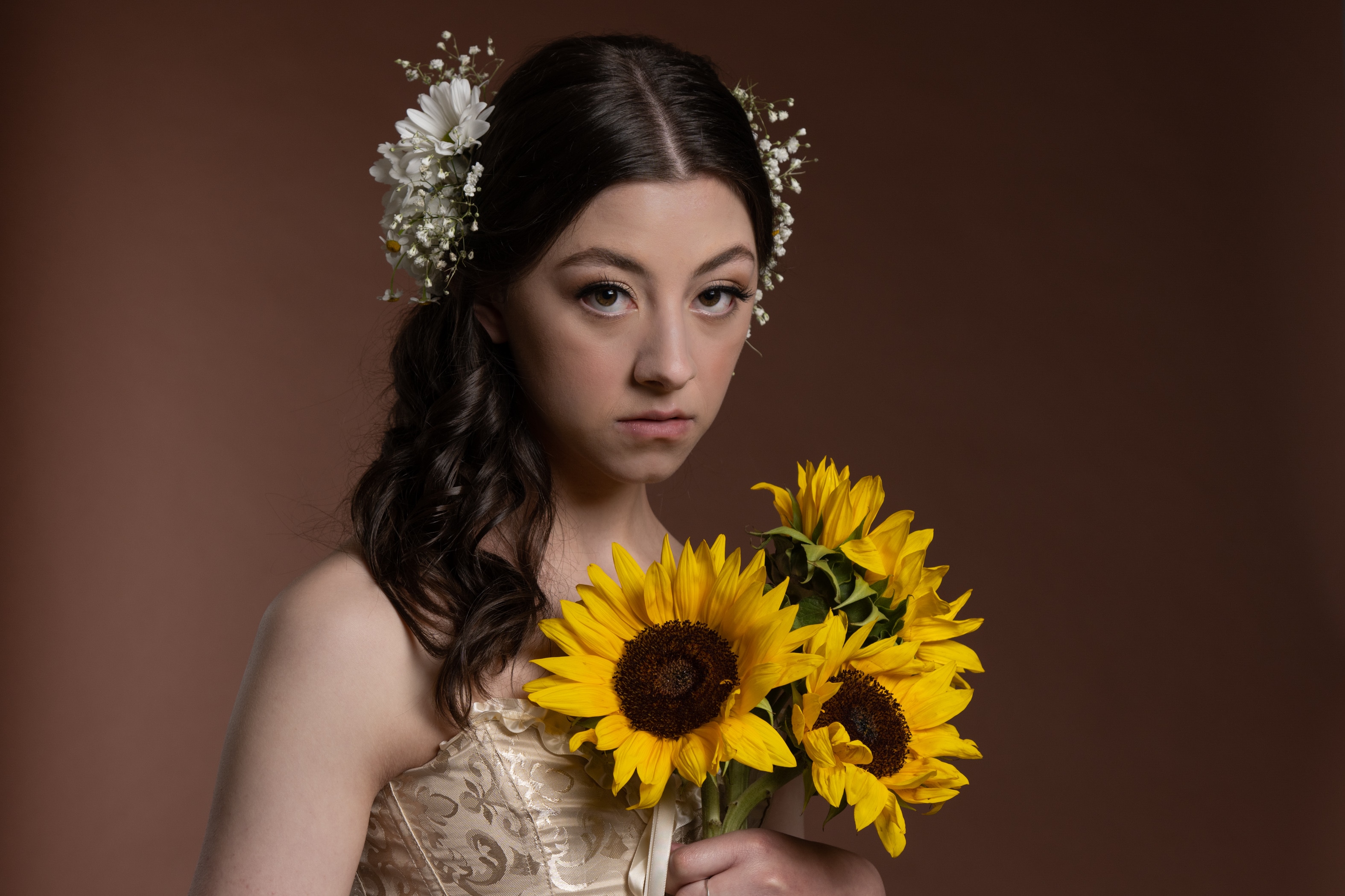 Young lady holding daisies with flowers in her long dark hair wearing a shoulder less white dress.