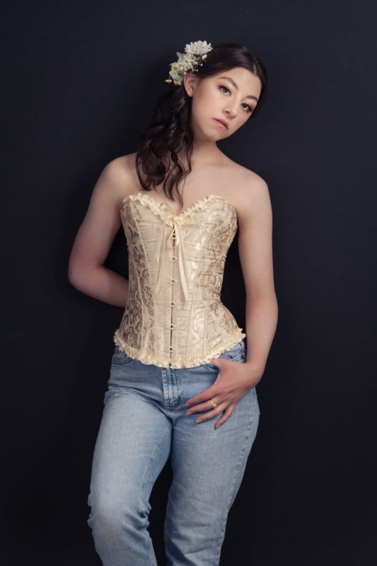Young woman posing for a portrait dressed in jeans with flowers in hair while leaning against a dark wall background.