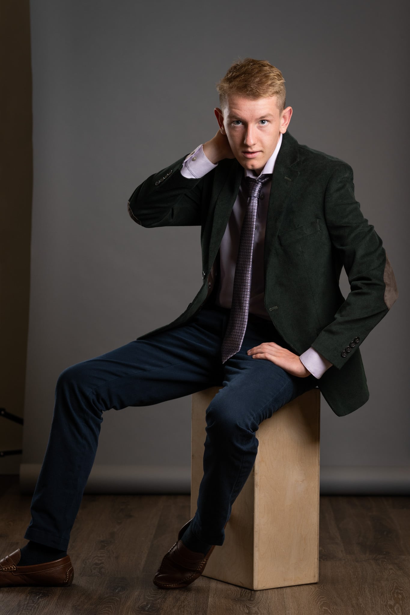 a high school senior guy sitting on a block stool wearing a tie, blazer, slacks, and posing