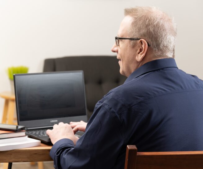 Portrait-Personal Branding photo of a man in a black fresh shirt sitting down and working on a black laptop at a desk.