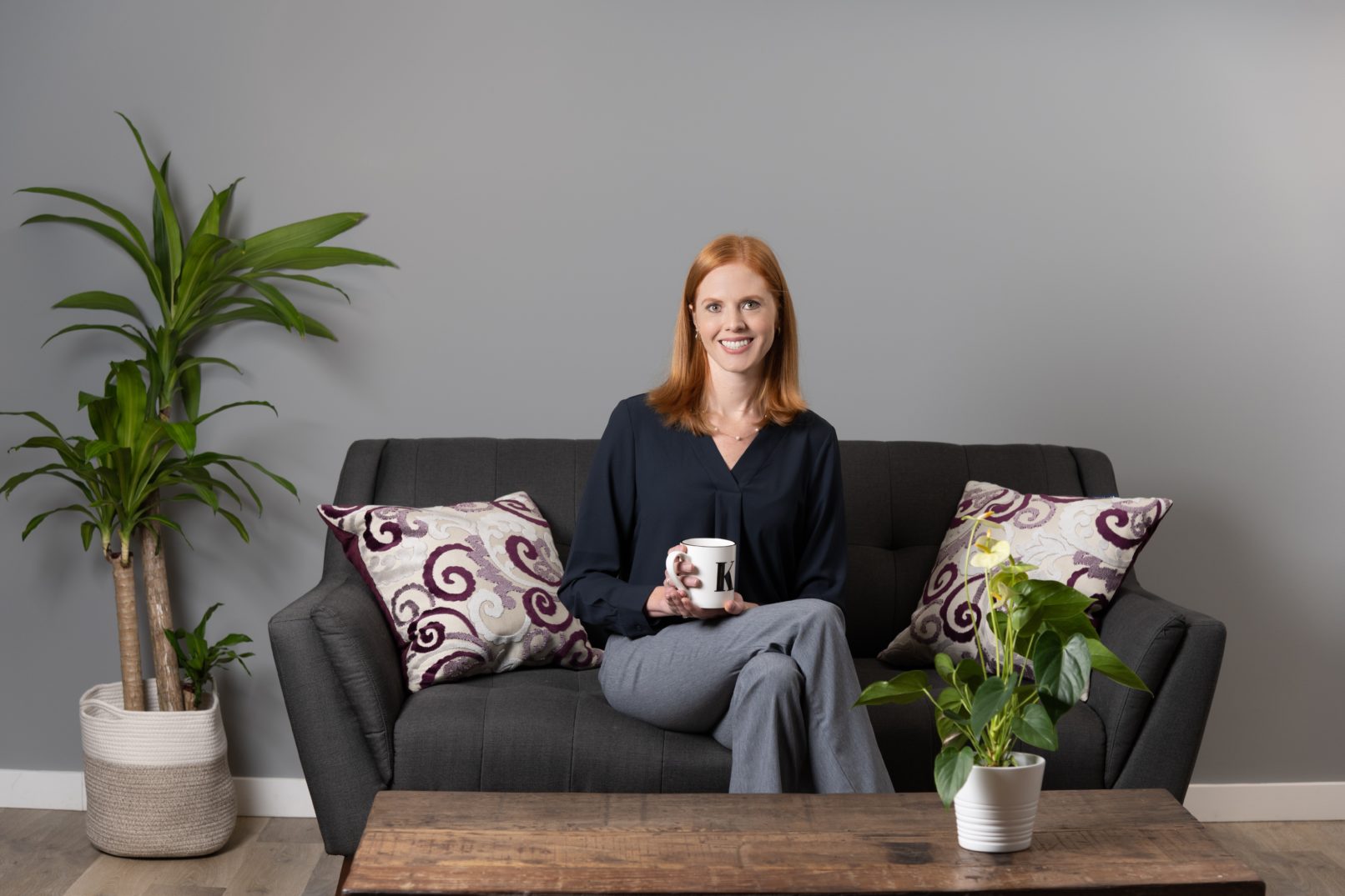Portrait-Personal Branding photo of a redhead woman sitting on a black couch with two pillows while she holds a white mug with the letter K printed on it in black.