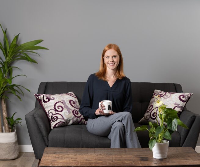 Portrait-Personal Branding photo of a redhead woman sitting on a black couch with two pillows while she holds a white mug with the letter K printed on it in black.