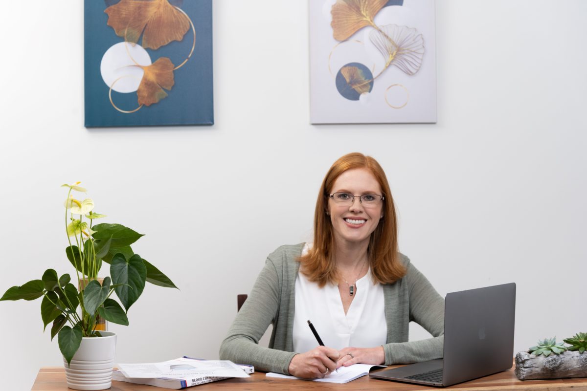 Portrait-Personal Branding photo of a redhead woman sitting at a desk and writing in a notebook while smiling towards the camera.