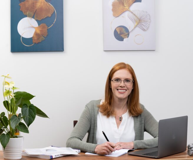 Portrait-Personal Branding photo of a redhead woman sitting at a desk and writing in a notebook while smiling towards the camera.