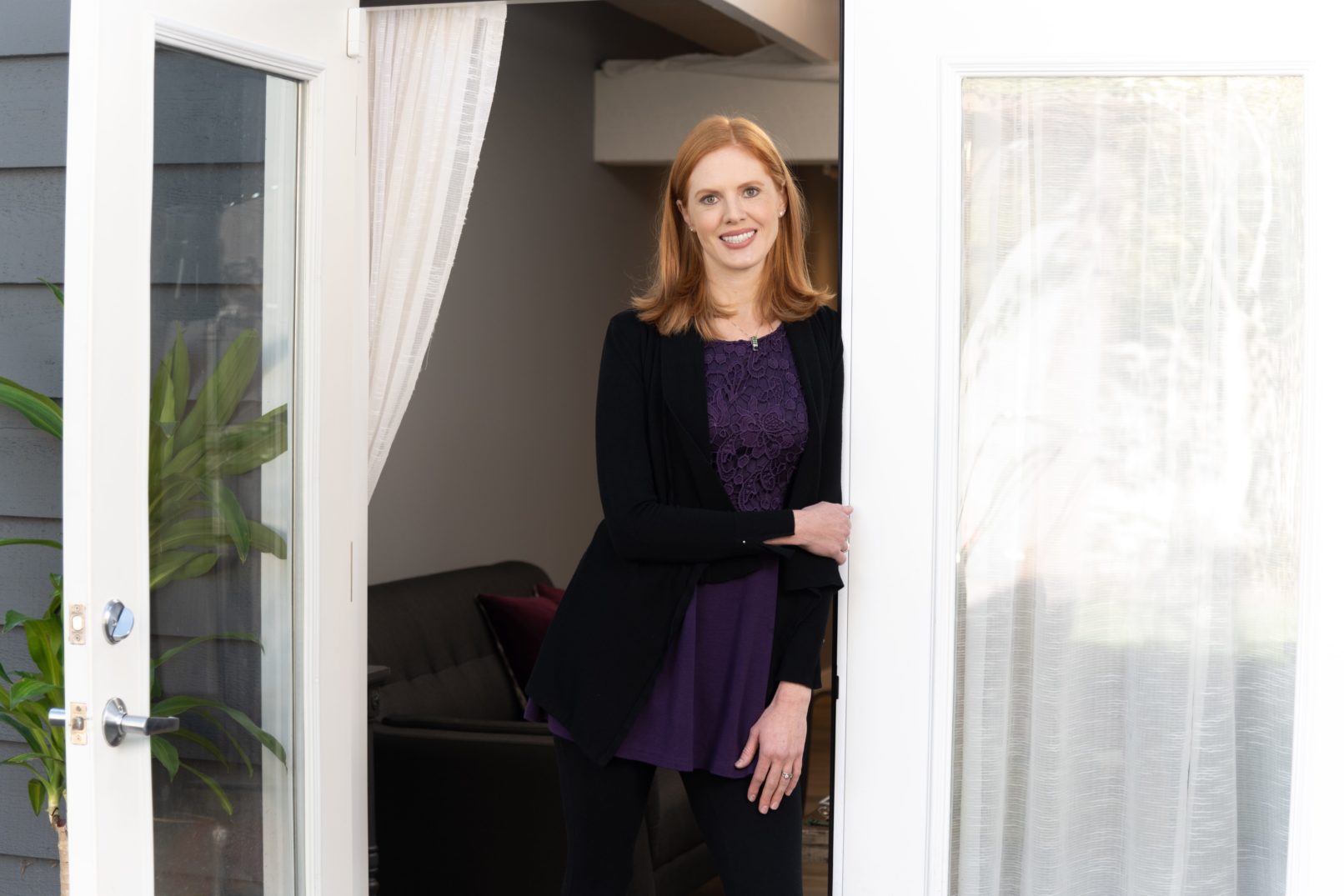 Portrait-Personal Branding photo of a redhead woman wearing a purple undershirt with a black sweater leaning in an open white doorway of a home.