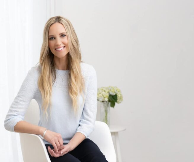 Portrait-Personal Branding photo of a blonde woman in a white sweater sitting down in a white chair inside of a white room with green flowers on a table behind her.