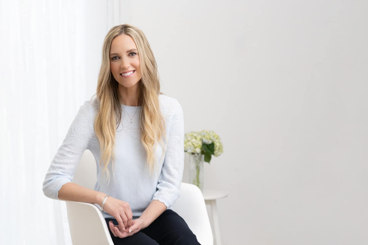 Portrait-Personal Branding photo of a blonde woman in a white sweater sitting down in a white chair inside of a white room with green flowers on a table behind her.