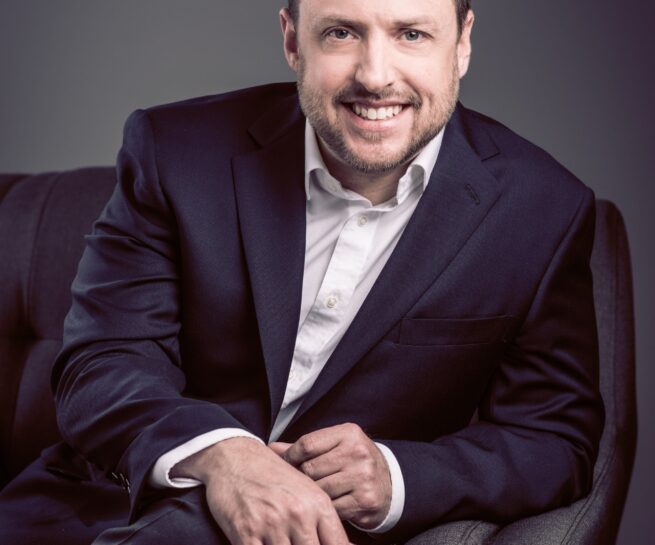 Portrait-Personal Branding photo of a brunette male in a black suit with no tie sitting on a grey couch while smiling towards the camera.