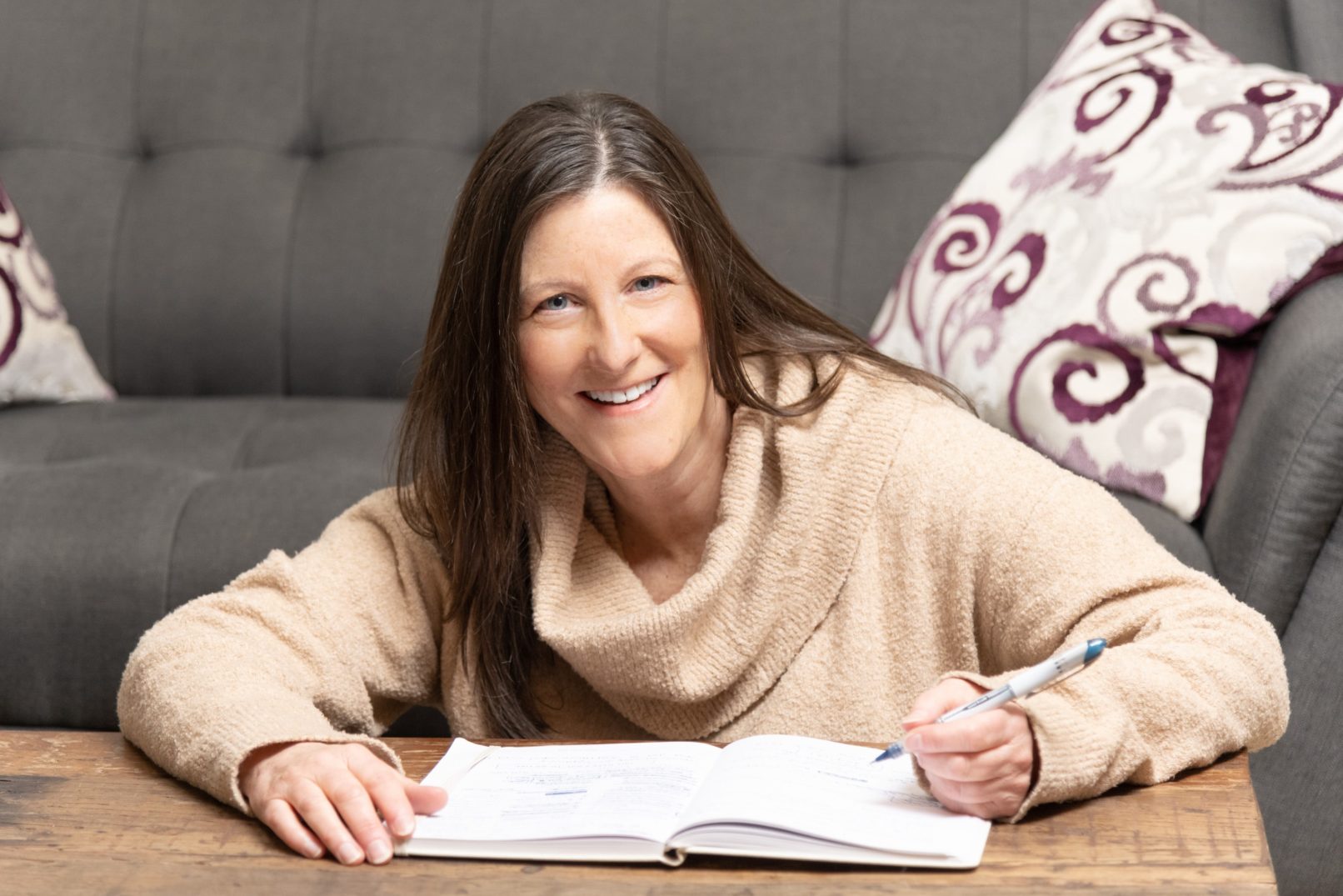 Portrait-Personal Branding photo of a woman smiling while sitting on floor at a coffee table writing in a book with a couch in the background.
