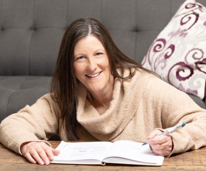 Portrait-Personal Branding photo of a woman smiling while sitting on floor at a coffee table writing in a book with a couch in the background.