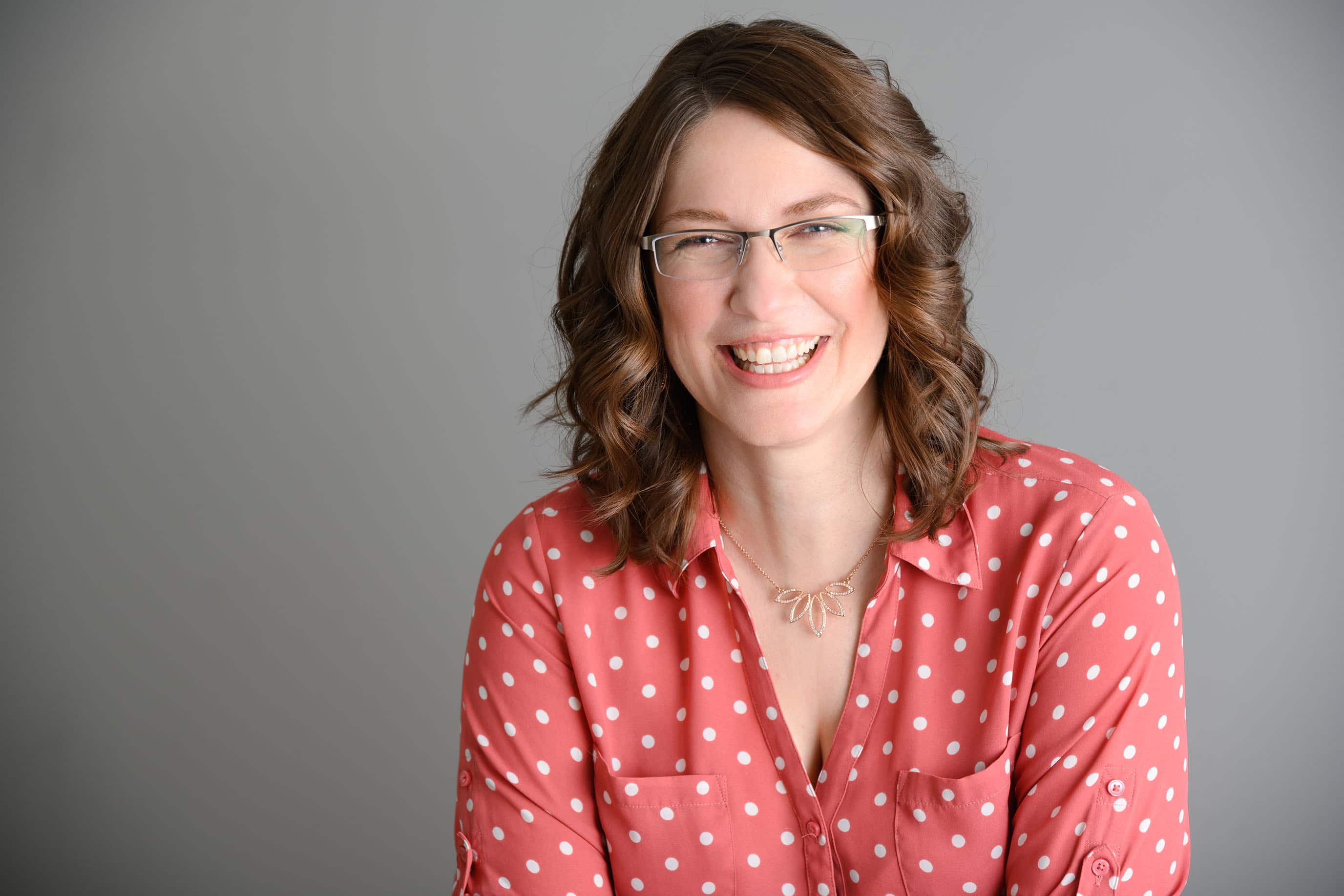 Headshot of a brunette woman with glasses in a red button-down polka dot shirt