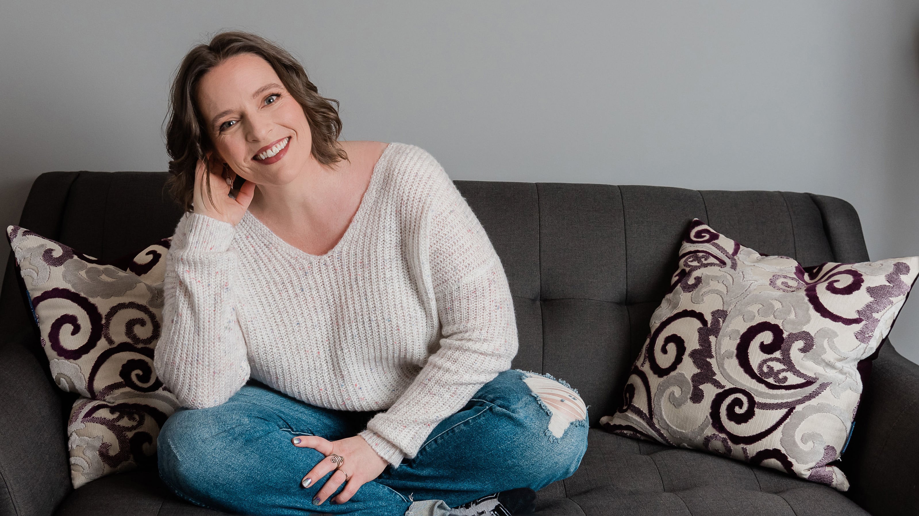Woman sitting on a charcoal grey couch, smiling wearing blue jeans and a white sweater, sitting legs crossed with feet on couch.