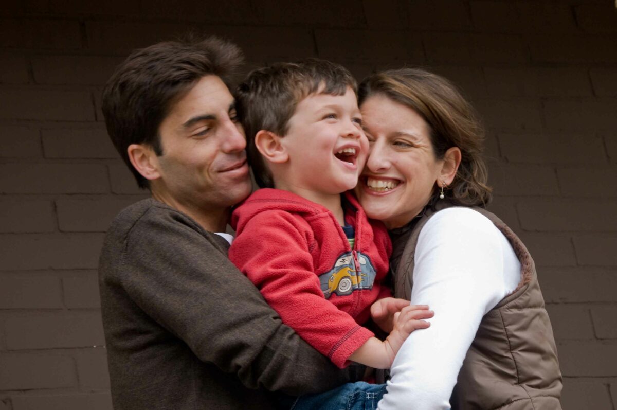 A mother and father pick up their young child and hug them together while laughing
