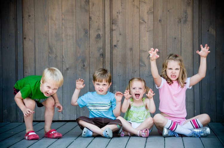 Four young children sit along a wooden floor and wall as they raise their hands like claws and make scary faces