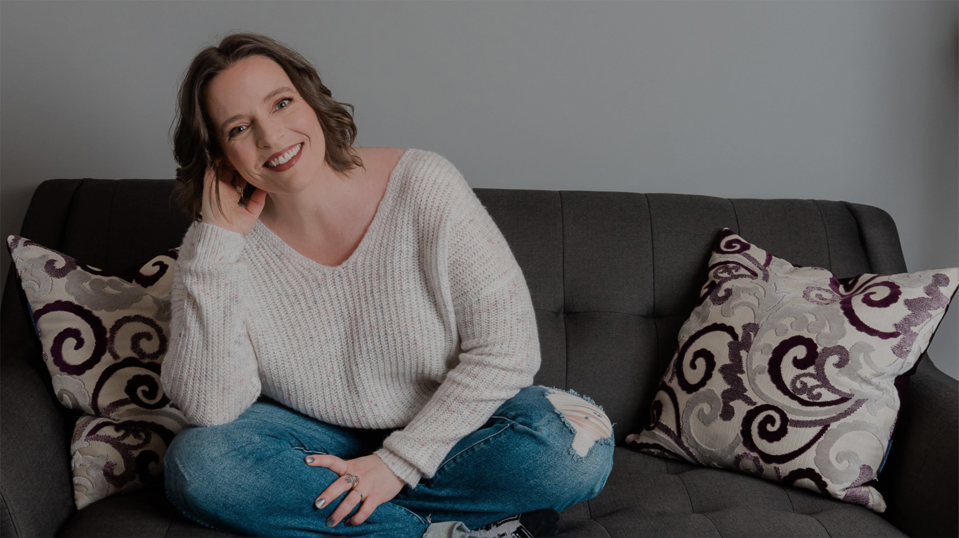 Middle aged woman casualy sitting on a dark colored sofa with two decrative pillows. Wearing faded jeans and oversized sweater and smiling.