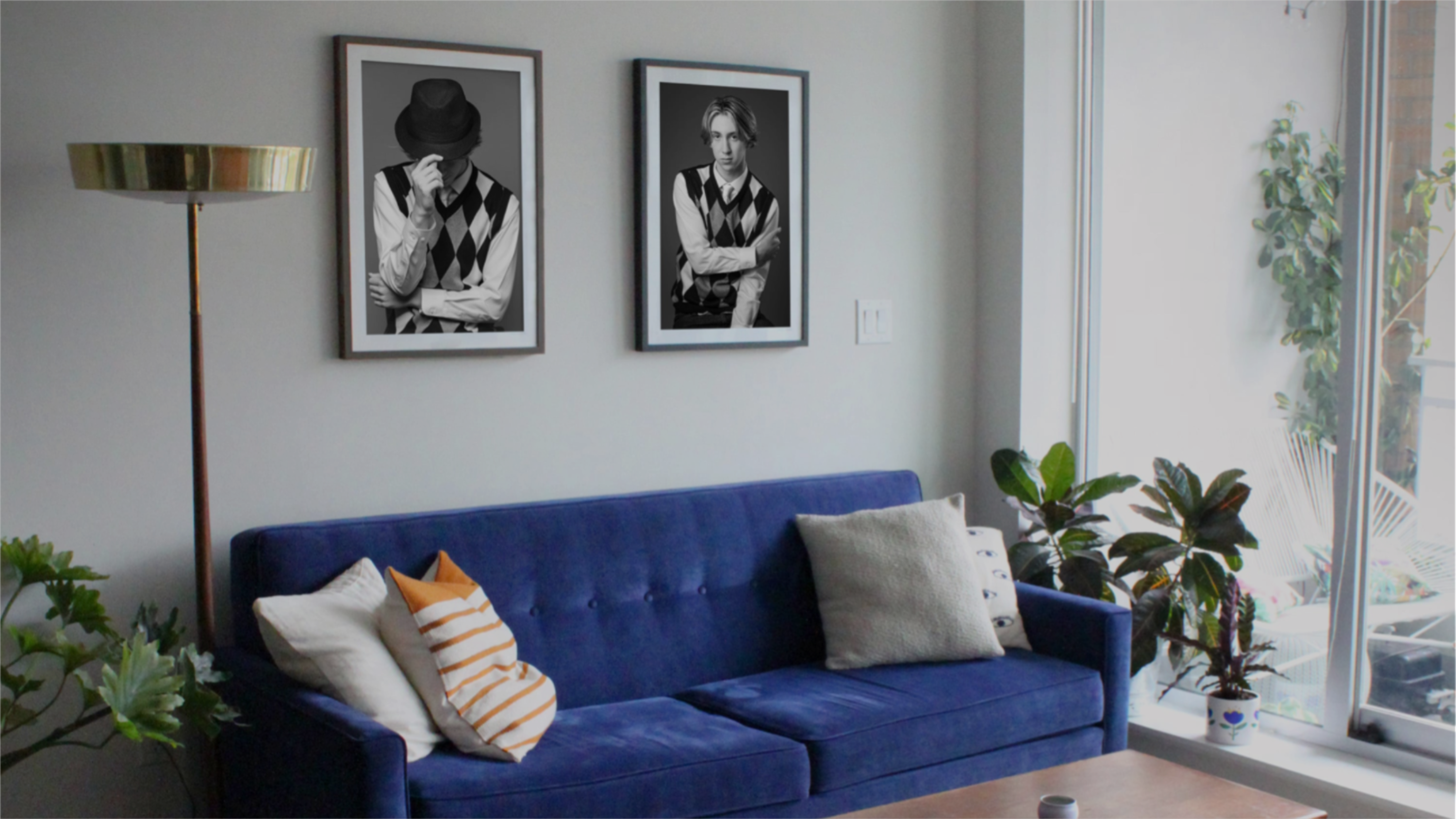Corner view of a n interior space, wall art, blue couch with four pillows plants, coffee table, floor to ceiling windows looking out on an outdoor space with chair and plant.