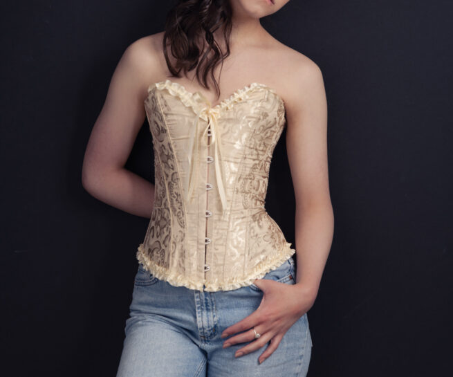High School senior Girl wearing a corset and jeans leaning against dark wall.