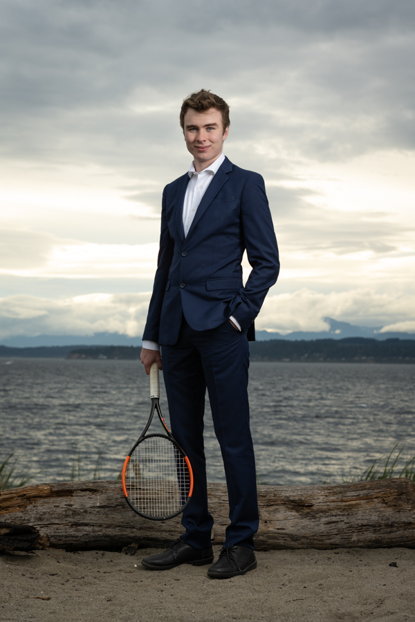 Man wearing an open shirt with blazer and dress pants, holding a tennis racket with left hand inside front pocket. Body of water and shoreline in background.