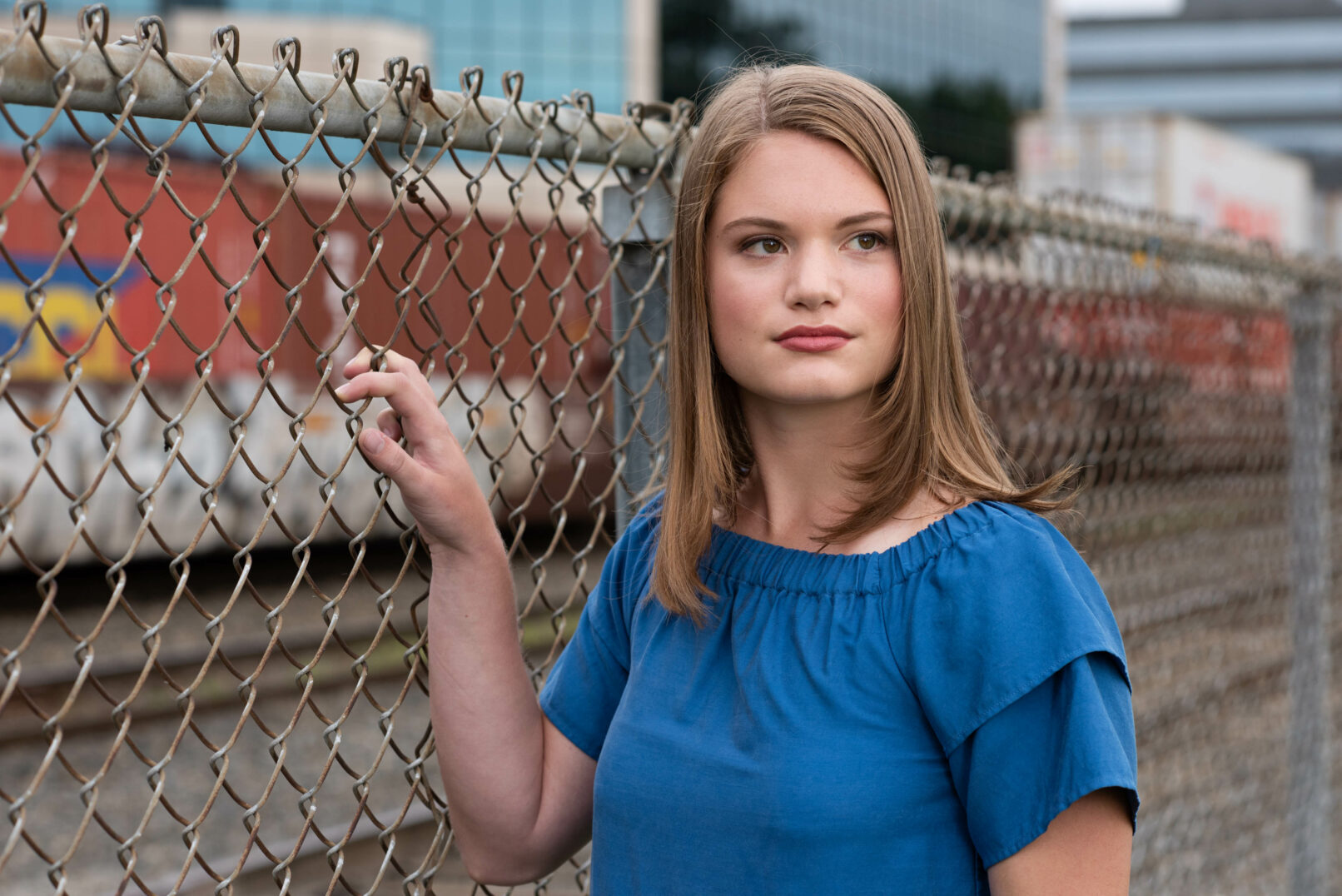young woman headshot standing next to chain link fence wearing a blue top. Train and office buildings in background.