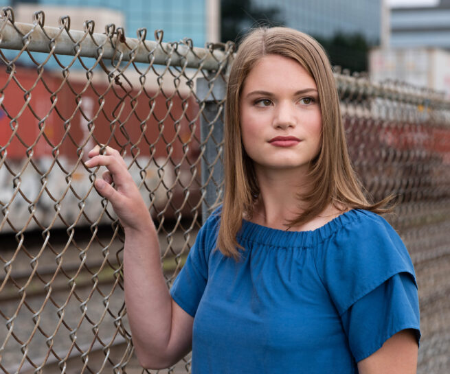 young woman headshot standing next to chain link fence wearing a blue top. Train and office buildings in background.